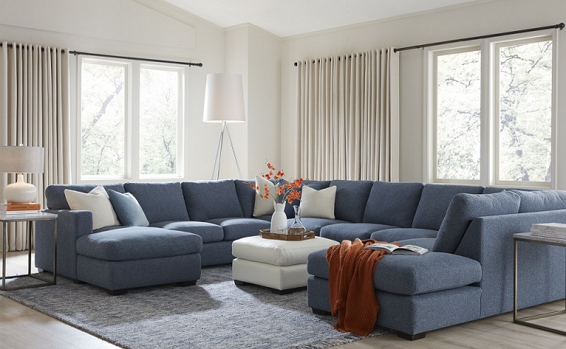 A living room with a greyish-blue sectional centred with white cushions. Two large windows on either side of the sectional in the background