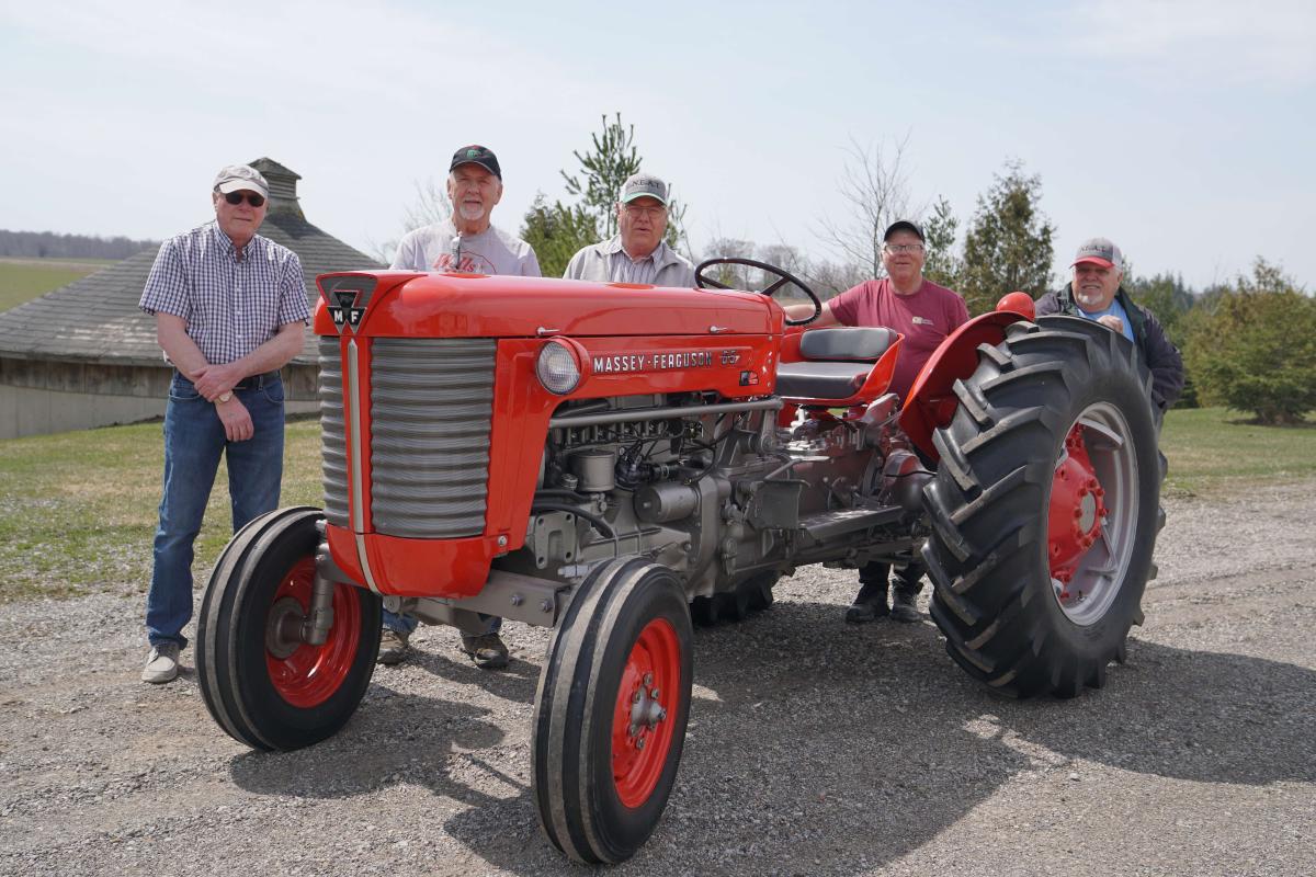 Five men with ball caps stand behind a red tractor with two large rear wheels and two small front wheels