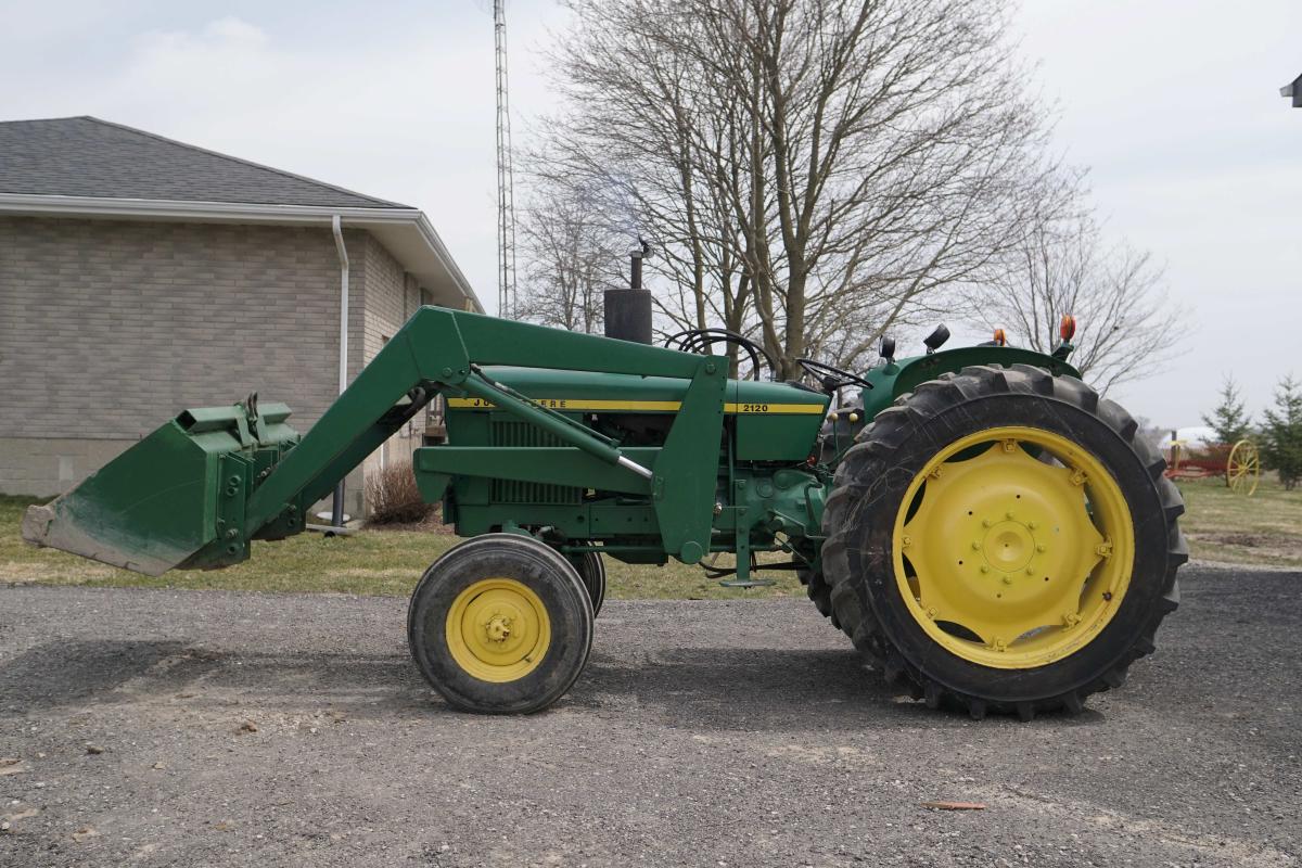 A side view of a green John Deere tractor with yellow rims. The plough is raised a few feet off the ground and a house and tree are in the background.