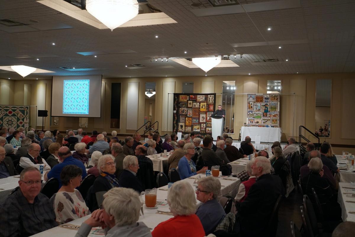 A man on stage overlooks a crowd gathered at tables to eat.