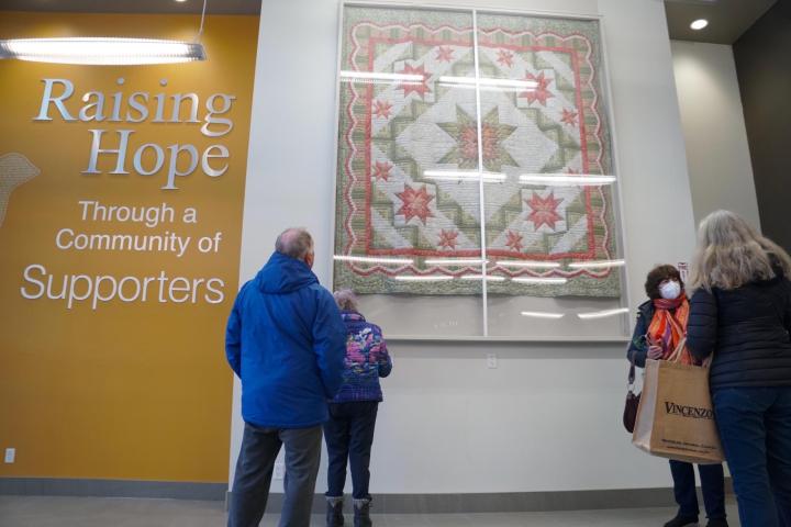 Three individuals look up at a quilt behind a protective cover hanging up high on a wall.