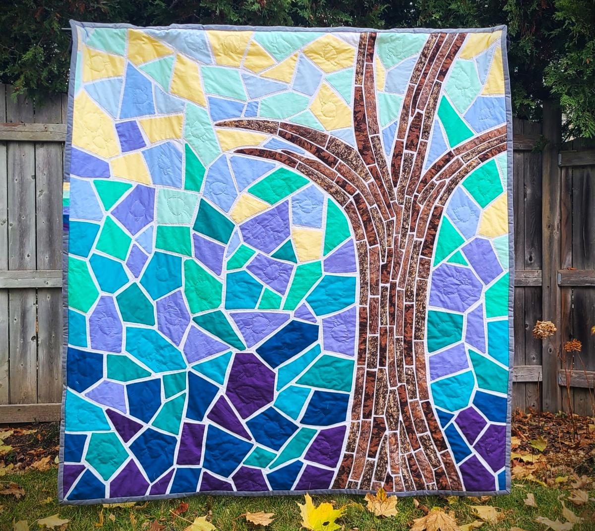 An image of quilt hanging outside in a garden with a fence behind it. The quilt is of a tree with a yellow and blue background.